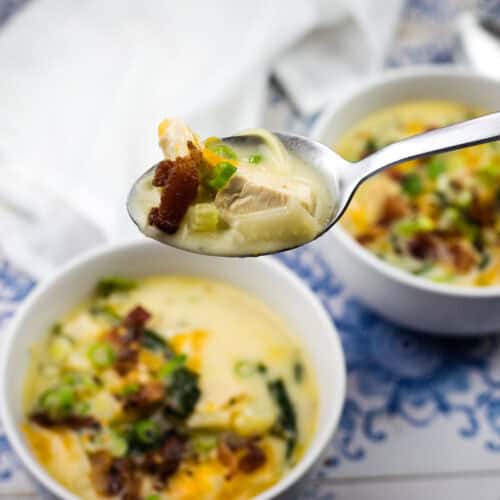 A close-up of a spoonful of creamy soup with chicken, bacon, and green onions held above two bowls of similar soup on a patterned surface.