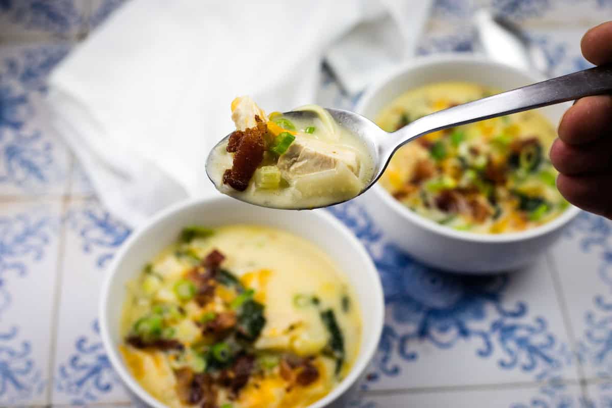 A close-up of a spoonful of creamy soup with chicken, bacon, and green onions held above two bowls of similar soup on a patterned surface.