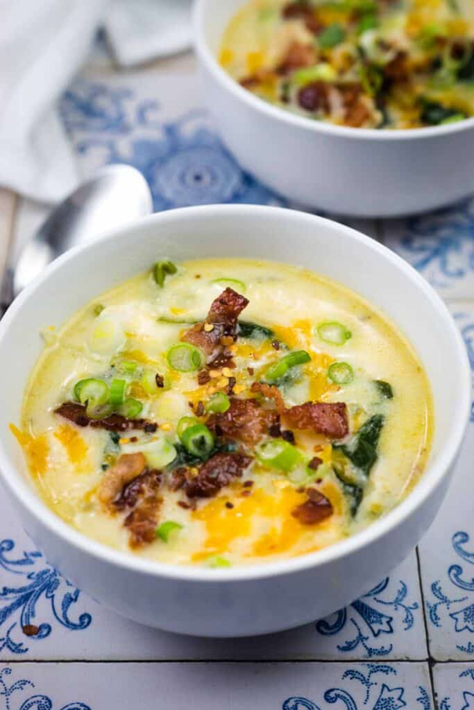 A bowl of creamy soup topped with chopped bacon, shredded cheese, and sliced green onions, with a spoon and a second bowl in the background.